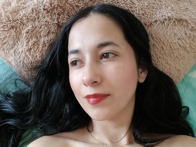 AnneStone's face, with long black hair, brown eyes, and red lips, lying on a fluffy pillow, showing a subtle smile, and wearing a thin necklace.