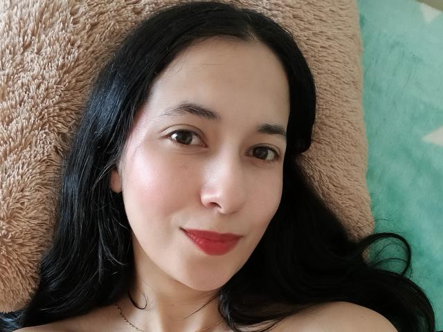 AnneStone has pale skin, black hair, brown eyes, red lipstick, and a slight smile, lying on a fluffy pillow with a green fabric behind her.
