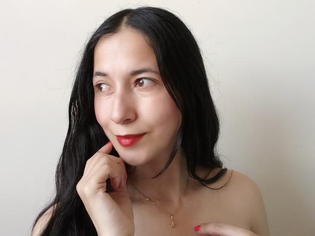 AnneStone with long black hair, fair skin, red lipstick, and a slight smile, touching her chin with her right hand, wearing a gold necklace, and showing pink nails.