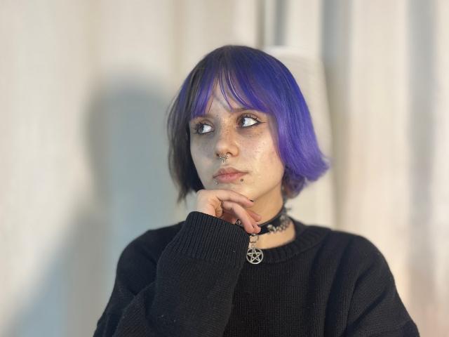 VickeyParkes with purple bob, black sweater, nose ring, and pentagram necklace, resting chin on hand, looking to the side, freckled face, soft light.