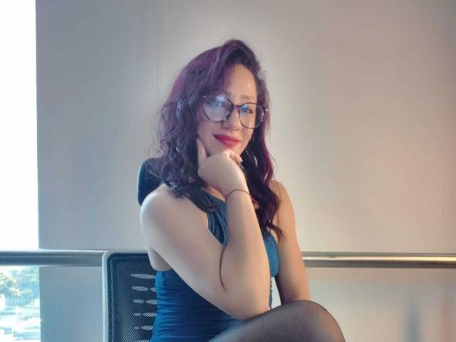 SamanthaCorine with curly purple hair, red lipstick, and glasses, in a blue dress, resting her chin on her hand, sitting on a metal chair, showing her ass.