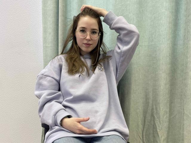 RobbieElsa mit Brille, helllila Kapuzenpullover und blauen Jeans, eine Hand in der Tasche, die andere im Haar, sitzt auf einem Stuhl, entspannt, mit einem grünen Vorhang im Hintergrund..