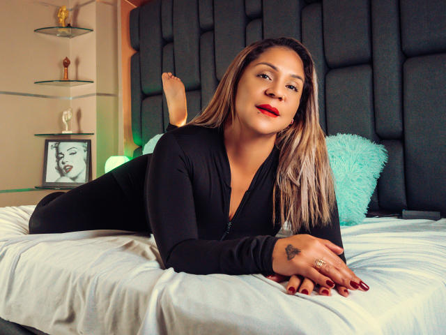 ManyHawkey, sdraiata sul letto con un top nero con zip, che mostra il sedere, con rossetto rosso, lunghi capelli biondi e un tatuaggio sulla mano destra, guarda la fotocamera, con i piedi sollevati, su un cuscino color turchese..