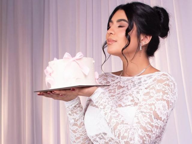 Antonella Montenblanc, con il seno coperto da una maglietta di pizzo bianco, tiene in mano una torta con un nastro rosa su un vassoio, con gli occhi chiusi, i capelli scuri raccolti in uno chignon e indossando degli orecchini..