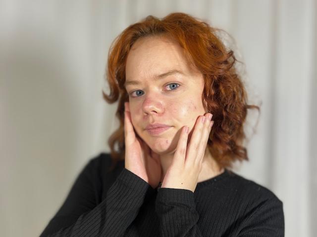 Corey Rounsaville a les cheveux roux et bouclés, des yeux bleus et une petite imperfection sur la joue. Elle porte un t-shirt noir côtelé, les mains sur le visage, et a l'air contrariée..