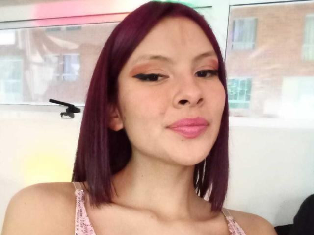 SamantaGreyy with purple hair, pink lipstick, and black eyeliner, pouting her lips. She's wearing a pink lace top, and has a small nose piercing. Her expression is playful.