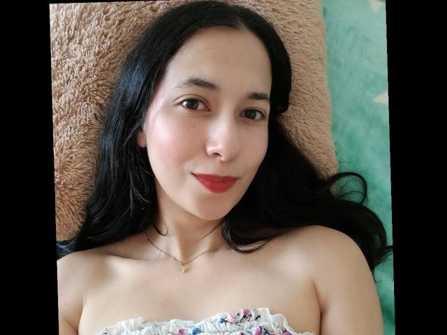 AnneStone with long black hair, light skin, red lipstick, and a subtle smile, lying down, wearing a strapless top, necklace, and no visible tits or ass.