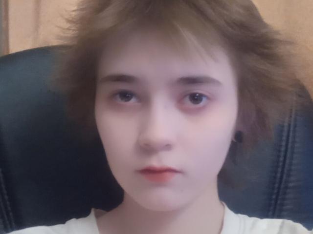 KarisaWinkel has pale skin, brown messy hair, blue eyes, small nose, and light pink lips. She's wearing a white shirt, sitting in a black leather chair.