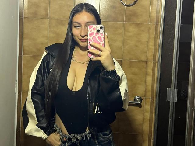 MaddyCarina, com cabelos castanhos longos, seios grandes, blusa preta, decote profundo, jaqueta preta e branca, segurando um celular rosa, banheiro com azulejos marrons, selfie no espelho, sorriso leve, visual casual..