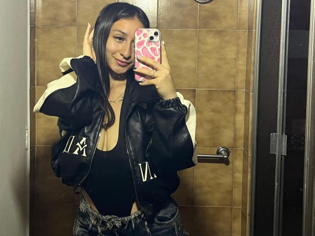 MaddyCarina with long black hair, wearing a black jacket with white stripes and "V" logos, smiling at the mirror, holding a pink phone, showing her tits in a black top.
