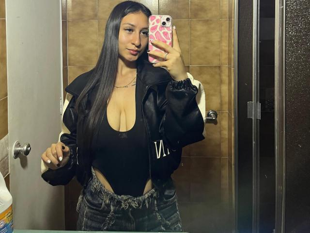 MaddyCarina, com longos cabelos pretos, vestindo uma jaqueta de couro preta, uma blusa preta decotada que mostra seus seios grandes e jeans de cintura alta, tirando uma selfie no espelho com um celular rosa..