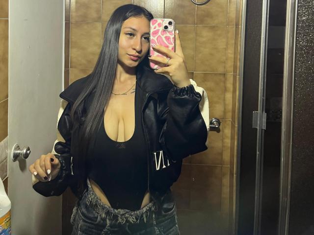 MaddyCarina with long black hair, wearing a black low-cut top showing her big tits, black jacket, and blue jeans, taking a mirror selfie in a bathroom.