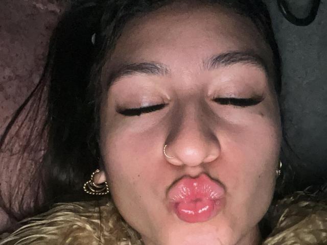 MaddyCarina's puffy ass-kissed face with eyes closed, nose ring, and gold earrings, showing her pink, shiny lips. She's got long dark hair and a cute, slightly sweaty look.