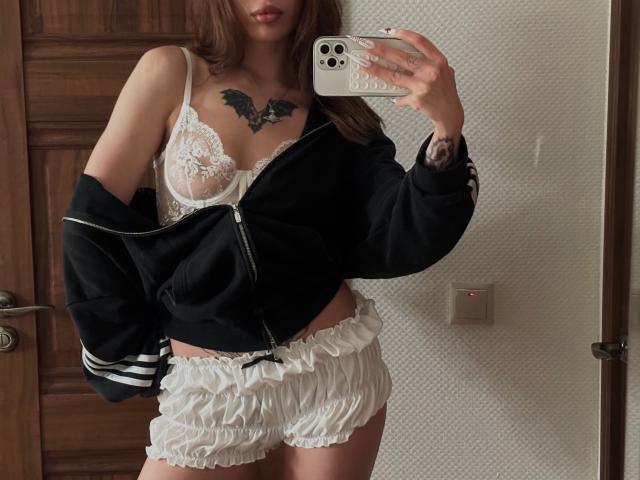 LilGingerMoon vestindo um sutiã de renda branco, mostrando seios pequenos, com uma jaqueta preta sem mangas, shorts brancos com babados, tatuagem no peito, segurando um celular, selfie no espelho, cabelo castanho, porta de madeira..