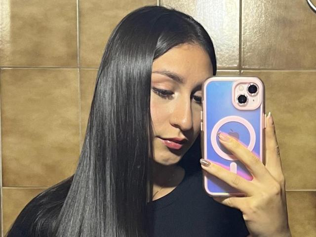 MaddyCarina with long black hair, holding a pink and blue phone, taking a selfie, showing her face and pink lipstick, wearing a black shirt, with manicured nails.