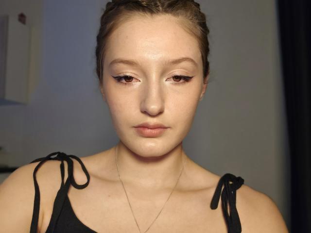 Close-up portrait of DiedraBloschichak with light skin and brown braided hair. She has subtle makeup with winged eyeliner and neutral lips. She wears a black, strap-tied top and a thin, gold necklace. Her expression is neutral, looking slightly downward. The lighting is even, highlighting her smooth skin and slightly shiny forehead. The background is blurred and indistinct. The image is clear with sharp details.