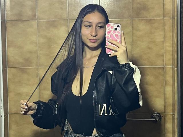 MaddyCarina with long black hair, wearing a black top and oversized black-and-white jacket, holding a phone with a pink heart case, pulling her hair. Brown-tiled bathroom.
