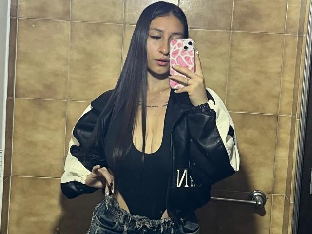 MaddyCarina with long black hair, wearing a black top with deep cleavage, black and white jacket, and high-waisted jeans, taking a mirror selfie with a pink phone.
