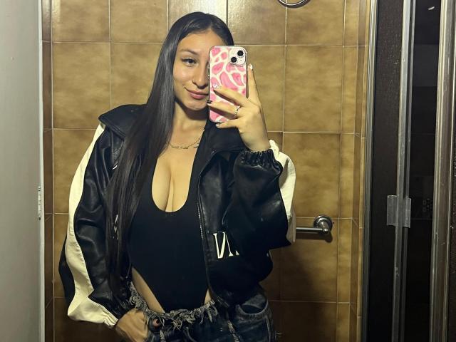 MaddyCarina met lang zwart haar, grote borsten in een zwarte top met diepe halslijn, een zwarte leren jas, die een roze telefoonhoes vasthoudt, staat in de badkamer en glimlacht naar een spiegelzelfportret..