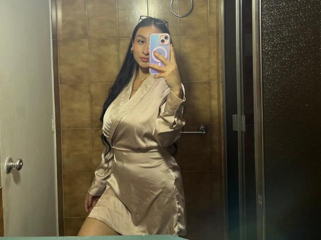 MaddyCarina with long black hair, wearing a silky beige dress, taking a mirror selfie, showing her tits, holding a phone with a pink pop socket. Bathroom setting.
