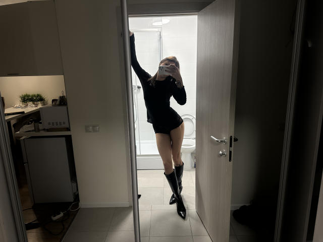 LilGingerMoon steht in einem Türrahmen, trägt ein schwarzes Langarmshirt, schwarze Shorts und knielange schwarze Stiefel und macht ein Selfie im Spiegel, wobei ein Arm angehoben ist..