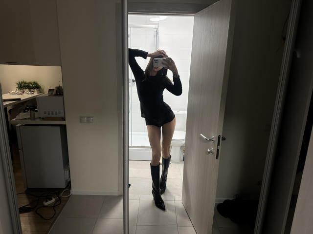 LilGingerMoon, com um corpo esguio e alto, vestida de preto, botas pretas e um celular cobrindo o rosto, está em uma porta, refletida em um espelho, mostrando suas nádegas..