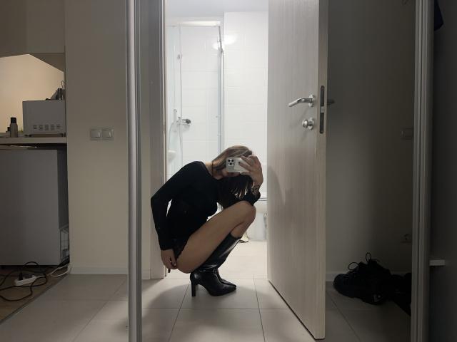 LilGingerMoon est accroupie devant un miroir, vêtue d'un haut noir à manches longues et de bottes noires à talons hauts, une main couvrant sa bouche. Elle est nue à partir de la taille..