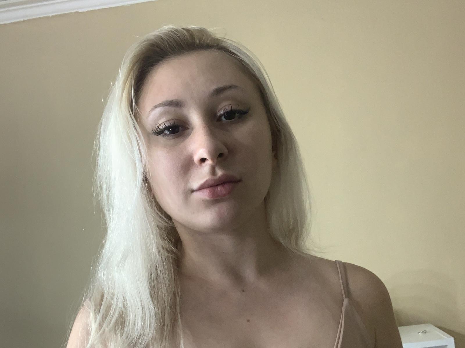 Helga Barragan, con i capelli biondi, indossa una canottiera beige, guarda in basso con un leggero sorriso, ha lunghe ciglia e un incarnato chiaro. Il suo seno è piccolo..