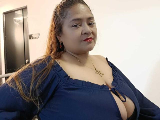 MonzerratBoyer with large tits, wearing a blue dress with a deep cleavage, long brown hair, red lipstick, gold necklace, and earrings, smiling in a simple indoor setting.