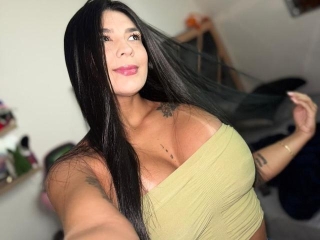 Antonella Montenblanc, con lunghi capelli neri, rossetto rosa, top verde che mette in mostra il suo seno prosperoso, tatuaggi sulla spalla e sul petto, che si tiene i capelli con la mano destra, sorridendo..