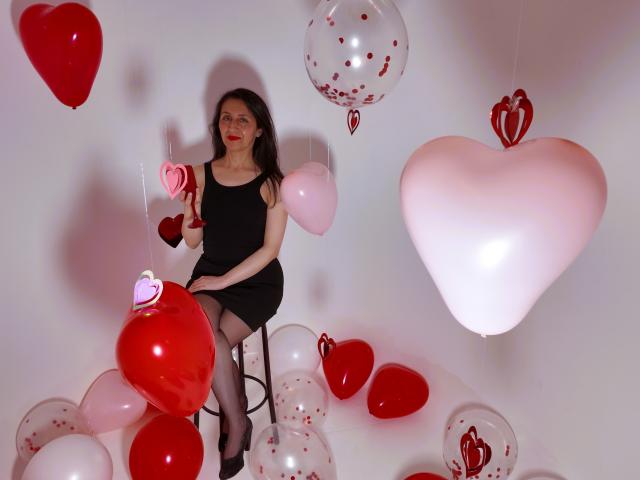 Cyntiia with tits, ass, and pussy-like balloon, sitting on stool, surrounded by red and white heart balloons, wearing black dress, stockings, and glasses, in a white room.