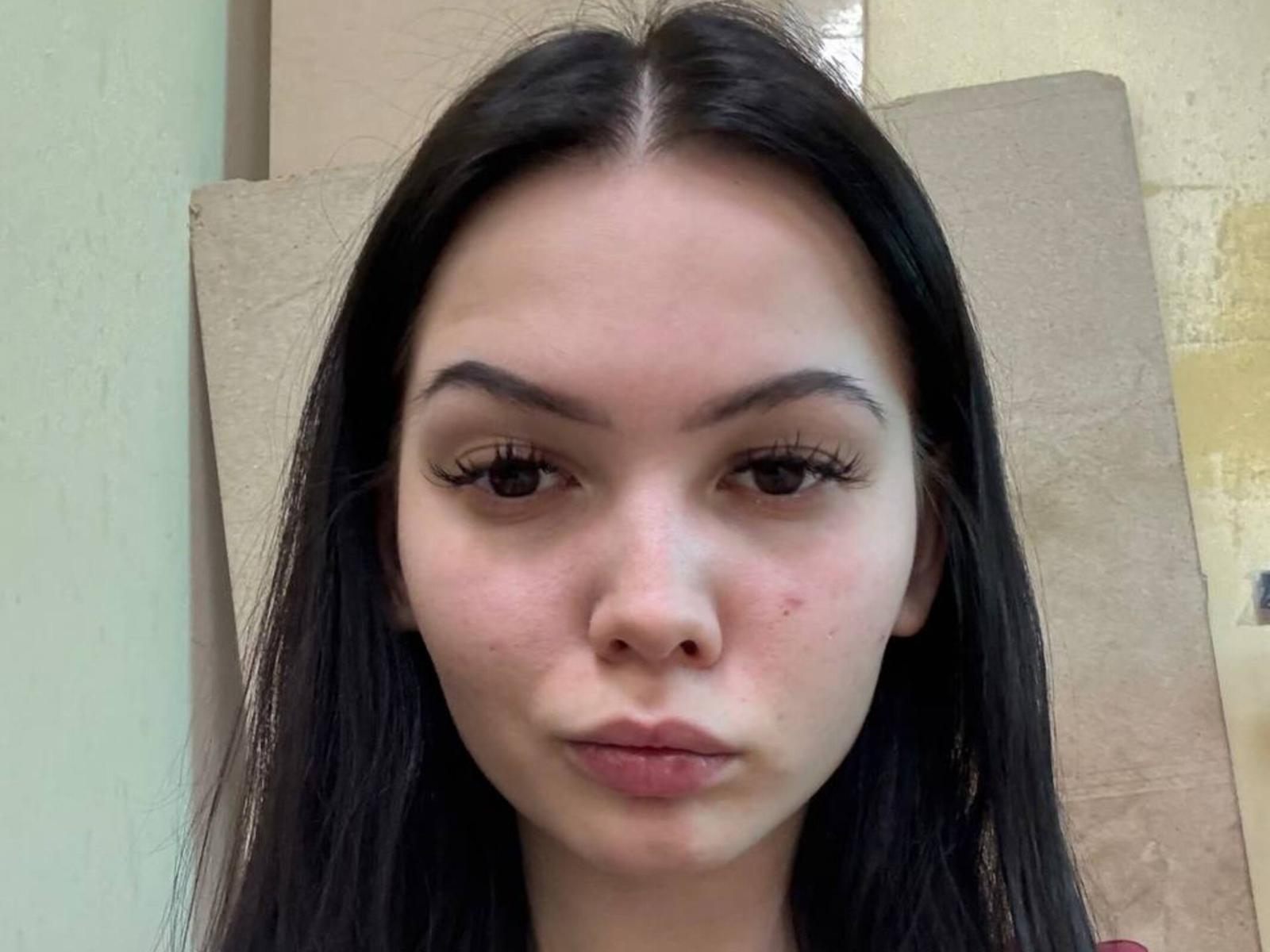CarrollGiorgianni has long black hair, brown eyes, and a small zit on her right cheek. She's looking straight at the camera with a neutral expression.