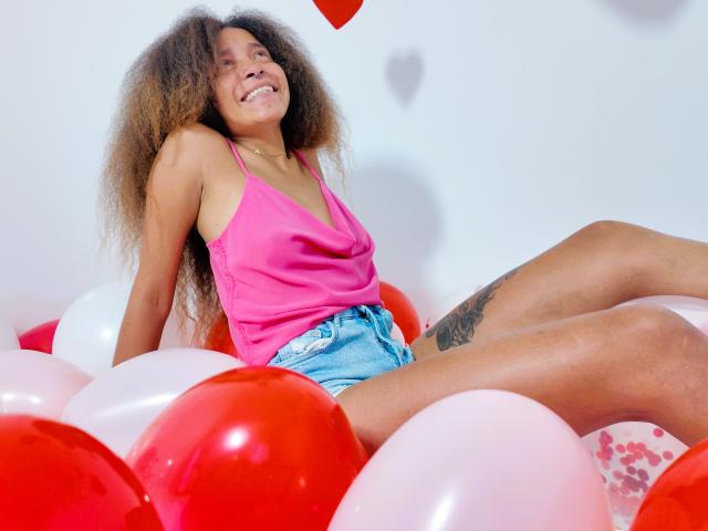 SweetSexx with curly brown hair, pink tank top, and denim shorts, sitting amidst red and white balloons, smiling, showing a tattoo on her right thigh.