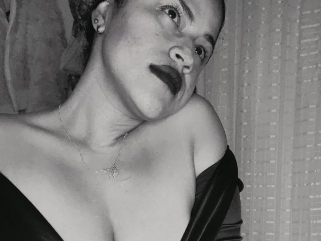 Black-and-white photo of IvannaDuran with dark lipstick, earrings, and necklace, leaning to the side, showing bare shoulders and small breasts, wearing a dark off-shoulder top.