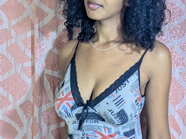 Ricka with curly black hair, medium brown skin, wearing a low-cut, patterned dress with black lace trim, showing her tits, and a silver necklace, sticking out her tongue.