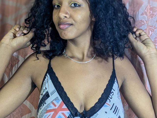 Ricka with curly black hair, wearing a low-cut, patterned top with black lace, exposing her small, brown, perky tits. She's smiling, hands in her hair, wearing a silver necklace.