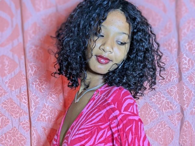 Ricka with curly black hair, red lipstick, and a pink patterned top, wearing a silver necklace, has a cute face and a slight smile. She's looking down.