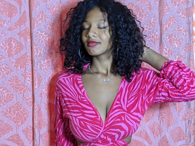 Ricka with curly black hair, pink zebra-patterned top, and pink lipstick, eyes closed, hand in hair, wearing a silver necklace, and large hoop earrings, standing against a pink patterned background.