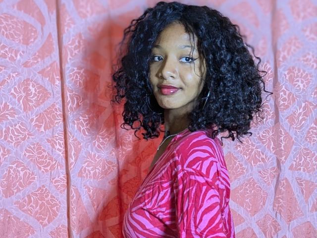 Ricka with curly black hair, wearing a pink zebra-pattern dress, red lipstick, standing against a pink floral curtain, looking over her shoulder with a subtle smile.