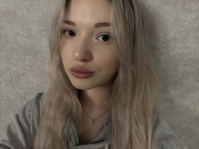 BulaBidgood with blonde hair, wearing a grey shirt, has full lips and a small necklace, her brown eyes staring forward, soft facial features, and a subtle, natural makeup look.