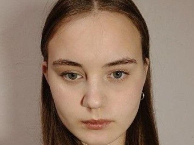 AlysiaFinkelstein has a fair, slightly pink complexion with long, straight brown hair parted in the middle. Her greenish-gray eyes are slightly downcast, and her full, slightly pursed lips are a muted pink. She has thin eyebrows and no visible makeup. She wears a small silver hoop earring on the left. The image is a close-up portrait with a neutral, slightly textured background. Her expression is neutral, almost somber.