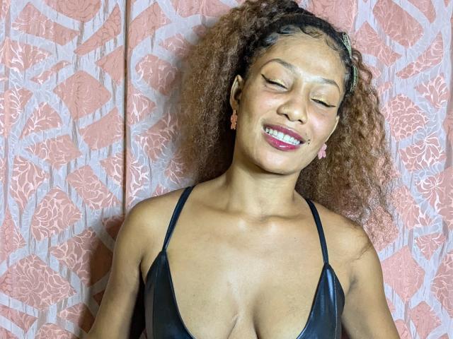 ClaraWish with curly brown hair, closed eyes, and pink lipstick, wearing a black, low-cut top, showing her tits. She's smiling, wearing pink earrings, against a pink patterned wall.