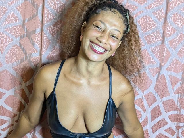 ClaraWish with curly brown hair, smiling, wearing a black leather bra, showing her tits, standing against a pink patterned curtain, looking happy and confident.