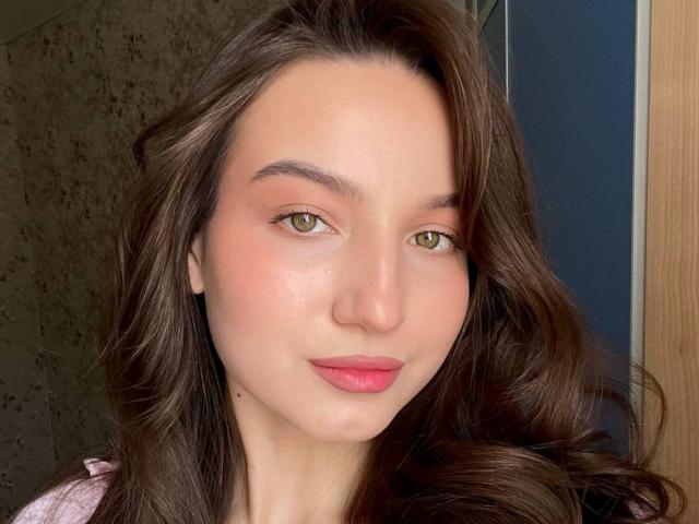 Margert Lewi heeft lang, bruin, golvend haar, groene ogen, roze lippenstift en een lichtrode huid. Ze draagt een roze top. Haar kleine neus en subtiele glimlach zijn zichtbaar..