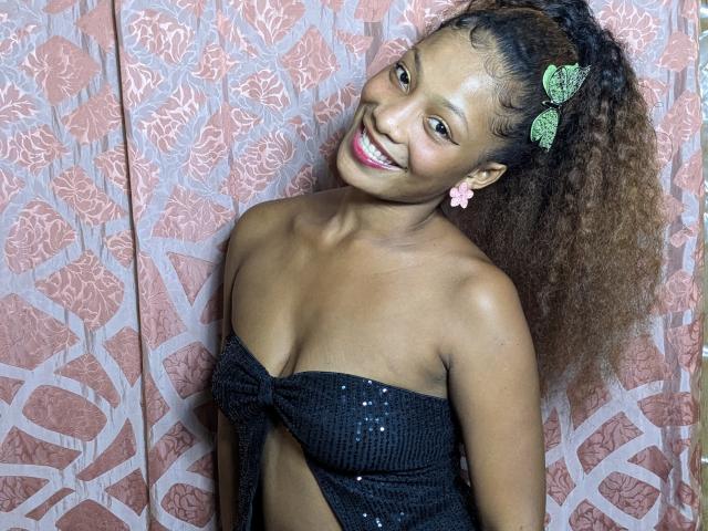 ClaraWish with curly brown hair, green butterfly hair clips, pink flower earrings, smiling, wearing a black sparkly strapless top, exposing her small tits, standing against pink patterned background.