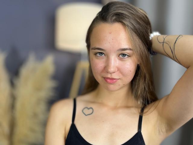 NickieZadd with straight brown hair, blue eyes, small heart tattoo on left chest, black tank top, arm raised, soft smile, slight acne, white wristband, blurry lamp in background.