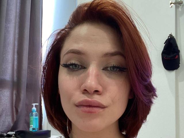 PeggyBehimer a les cheveux roux, les yeux verts, et un trait d'eye-liner noir avec de longs cils. Elle regarde l'appareil photo avec un léger sourire. Son visage est centré dans l'image..