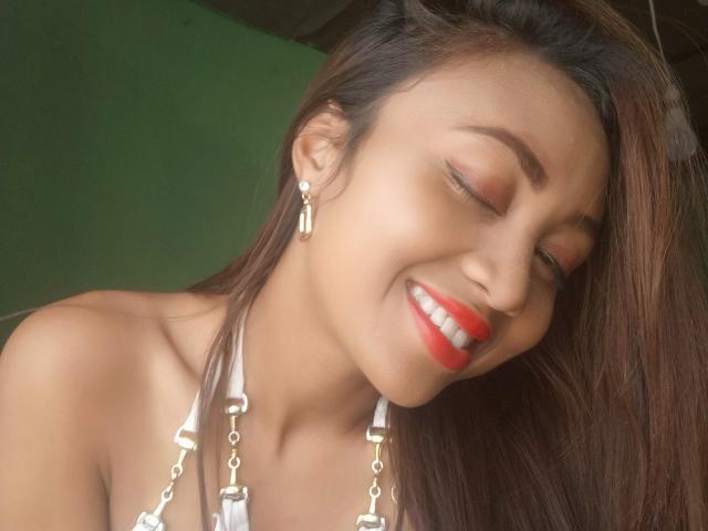 O rosto sorridente de Narami, com os olhos fechados, usando batom vermelho, um top branco de alças, brincos de prata, com cabelos castanhos longos, e uma expressão adorável, bonita, sexy, atraente e feliz..