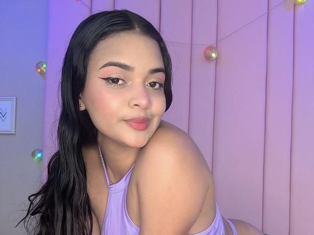 StacyReese with black hair, pink eyeliner, and purple top, showing her bare shoulder and puffy pink lips, sitting with a slight smile, looking at the camera.