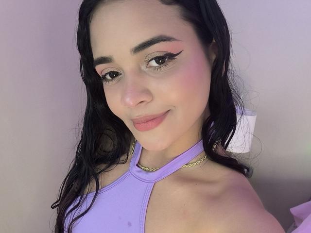 StacyReese with pink eyeshadow, black eyeliner, and a subtle smile, wearing a purple halter top and gold chain necklace, with wet black hair. Soft pink blush on her cheeks.